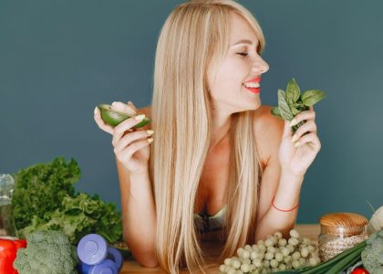 A smiling woman holding avocado and basil, surrounded by fresh vegetables and fruits for healthy eating.